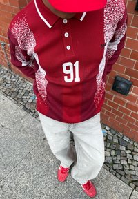 Rotes Poloshirt mit weißen Akzenten und der Nummer 91, kombiniert mit hellgrauer Hose und roten Turnschuhen. Strukturierter Stoff mit dynamischem Muster.