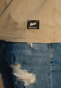 Beige bomulls t-skjorte med en svart etikett med hvit tekst; brukte lyseblå jeans med slitte rifter og frynser ved knærne.