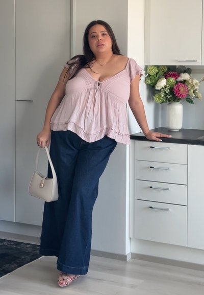 Mujer con blusa rosa y pantalones anchos azul marino de pie en una cocina moderna sosteniendo un pequeño bolso beige cerca de un jarrón con flores de colores.