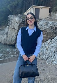 Femme portant des lunettes de soleil, chemise bleue avec un gilet noir et un jean, tenant un sac à main noir, debout sur une plage rocheuse près de l'eau et d'un petit bâtiment.