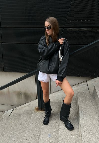 Chaqueta bomber de piel negra, pantalones cortos blancos con puntilla, botas negras hasta la rodilla con hebillas, bolso plateado, de pie en unas escaleras contra un fondo texturizado negro.