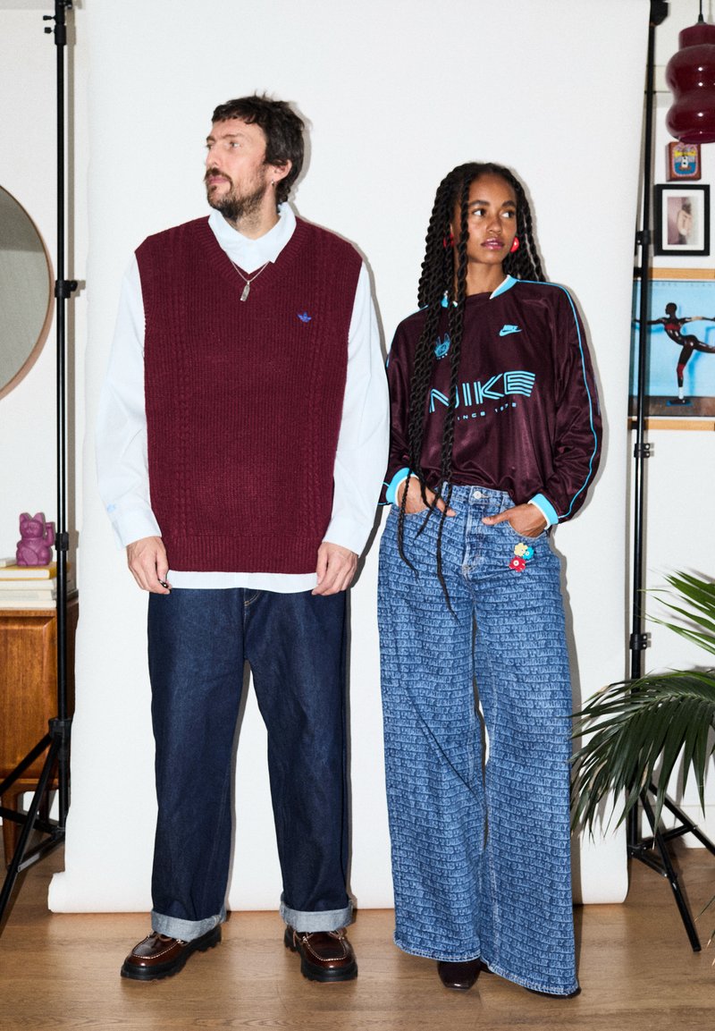 Άνδρας με μπορντό πλεκτό γιλέκο και λευκό πουκάμισο στέκεται δίπλα σε γυναίκα που φοράει σκούρο φούτερ Nike και φαρδιά τζιν με λουλουδάτα μπαλώματα.