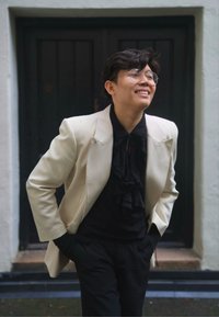 Kremfarget blazer over en svart skjorte med flæser, svarte bukser og runde briller. Glatt stoff med strukturert design, mot en mørk dør.