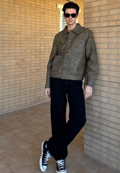 Chaqueta texturizada verde oliva con bolsillos delanteros, combinada con pantalones negros y zapatillas Converse negras, con un fondo de ladrillo.