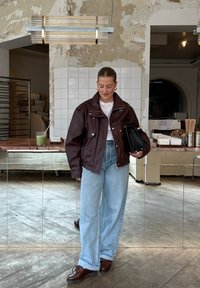 Donna che indossa una giacca di pelle bordeaux, jeans a gamba larga blu chiaro, scarpe marroni, mentre tiene una borsa nera in un caffè rustico con pavimenti in legno e pareti piastrellate.