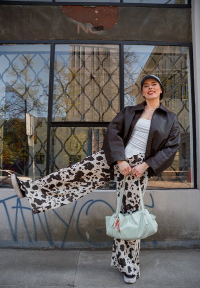 Joven mujer con pantalones de estampado de vaca, top blanco, chaqueta oscura y gorra posa con una pierna levantada, sosteniendo un bolso verde menta en una acera urbana.