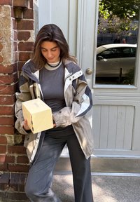 Giacca grigia chiara e grigia scura con dettagli bianchi, top grigio e pantaloni ampi grigi. Tiene in mano una scatola beige con un logo. Muro in mattoni e porta sullo sfondo.