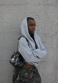 Haut à capuche gris avec une capuche, pantalon camouflé, et un sac noir brillant avec des anses en chaîne. L'arrière-plan présente un mur gris texturé.