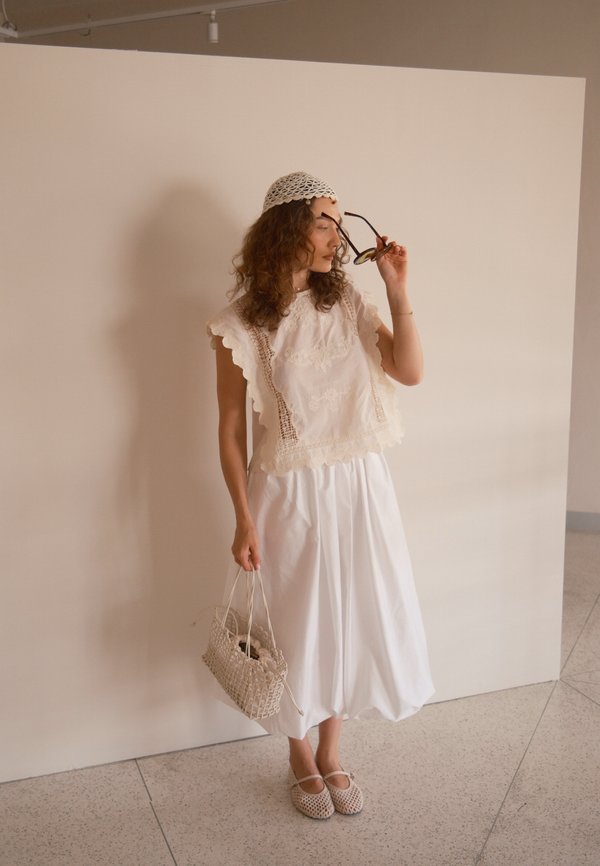Femme portant un haut en dentelle blanche, une jupe blanche, une casquette au crochet, tenant des lunettes de soleil et un sac tressé, se tenant contre un mur beige uni.