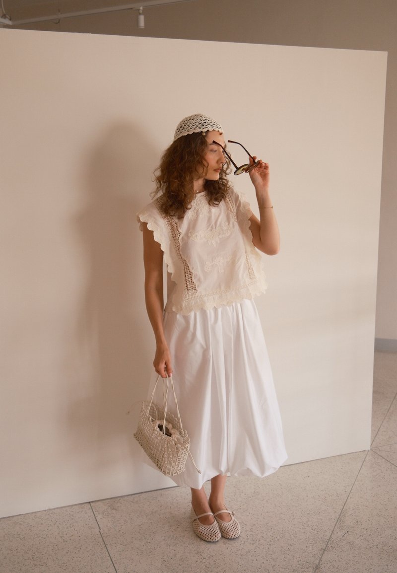 Femme portant un haut en dentelle blanche, une jupe blanche, une casquette au crochet, tenant des lunettes de soleil et un sac tressé, se tenant contre un mur beige uni.