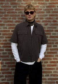 Hemd mit kurzen Ärmeln in Braun, über einem weißen Langarmshirt getragen, kombiniert mit dunklen Jeans und Sonnenbrille, vor einer Ziegelwand.