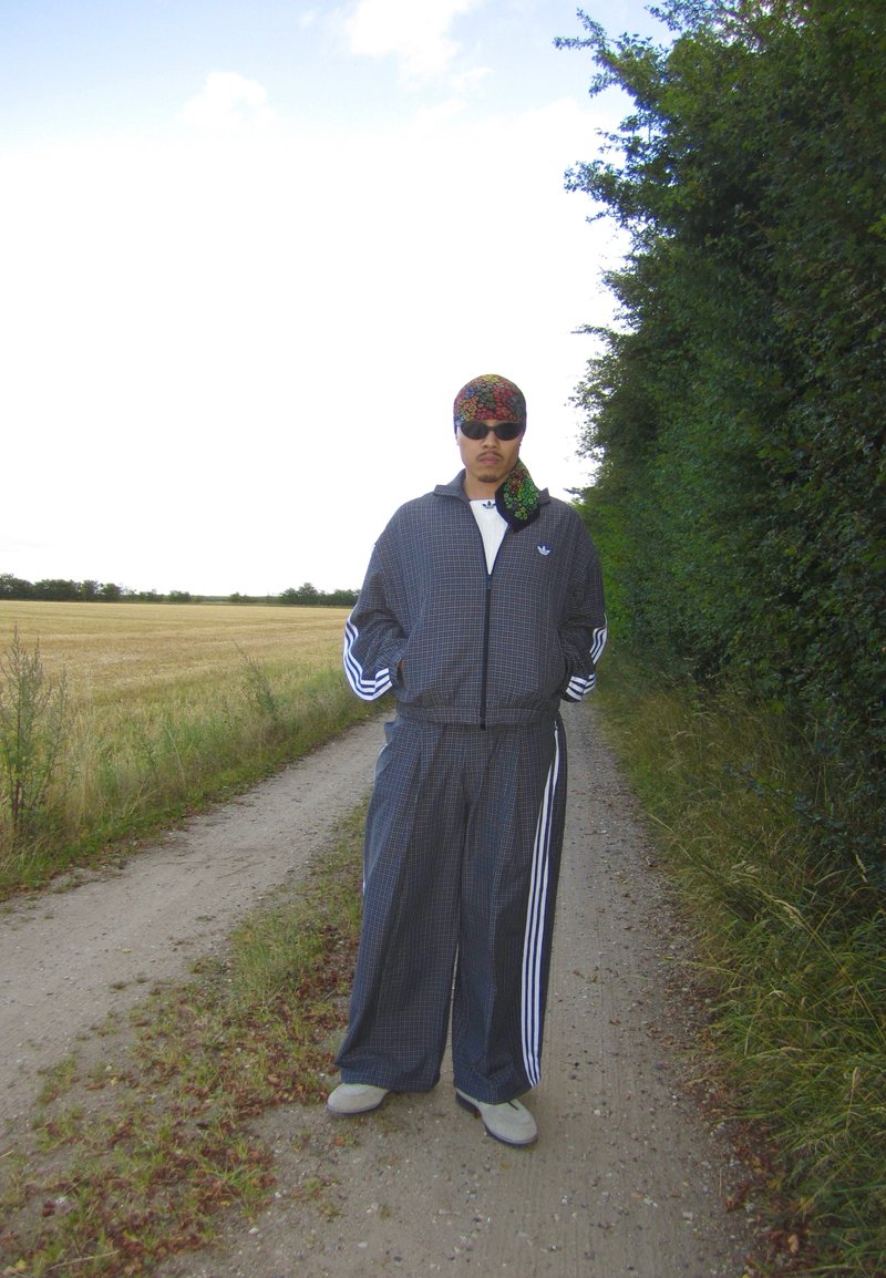 Personne portant un survêtement foncé, des lunettes de soleil et un foulard coloré, debout sur un chemin en terre entre un champ et des arbres.
