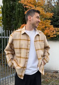 Camicia a quadri beige con linee gialle e marroni, maniche lunghe e design con bottoni, indossata sopra una t-shirt bianca a coste. Dotata di una tasca sul petto.