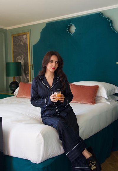 Pijamas de satén a rayas marinas con ribete blanco, sentados en una cama con sábanas blancas y cojines de acento melocotón, sosteniendo una bebida en un vaso.