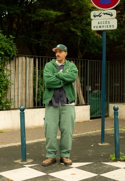 Sudadera verde y pantalones anchos verdes, combinados con una camiseta gris. Zapatos de estampado de leopardo. De pie cerca de un poste azul y una señal de tráfico.
