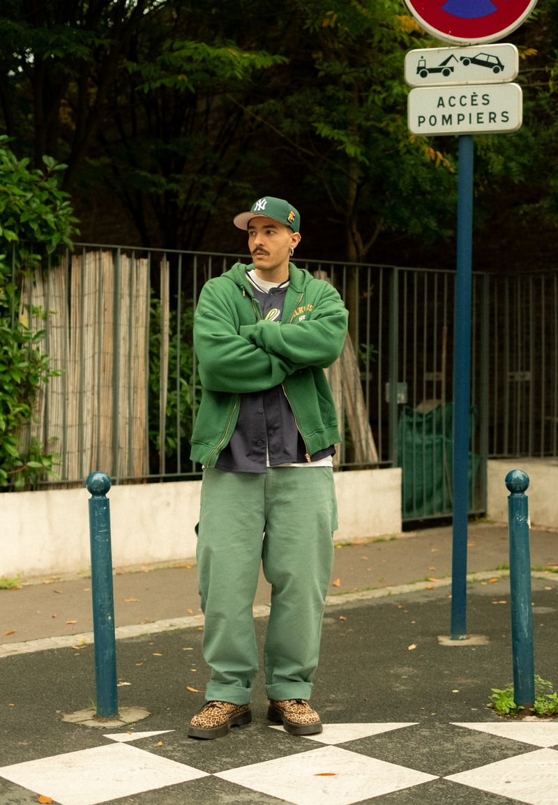 Moletom verde e calças largas verdes, usados com uma camisa cinza. Sapatos com padrão de leopardo. De pé perto de um poste azul e uma placa de trânsito.