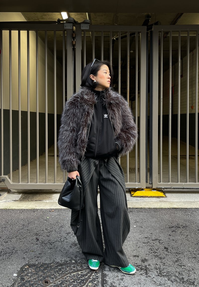 Femme en veste fausse fourrure sombre, haut Adidas noir, pantalon large à fines rayures, chaussures vertes, tenant un sac noir, debout près d'une porte métallique.