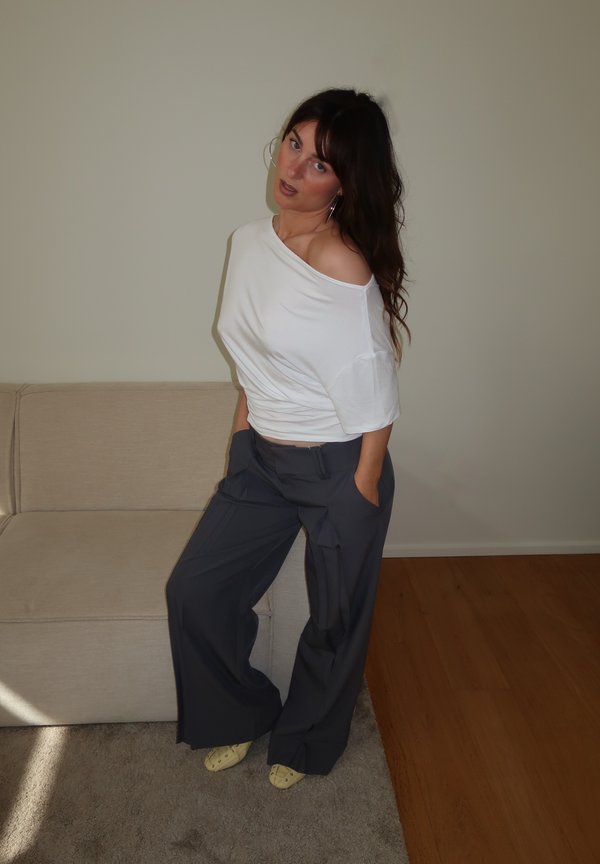 Kobieta o długich ciemnych włosach, ubrana w biały top z odkrytymi ramionami, ciemne szerokie spodnie i żółte buty, stojąca w pomieszczeniu obok beżowej kanapy.