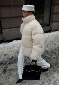 Femme marchant dans une rue enneigée, portant un manteau blanc duveteux, un pantalon blanc, un chapeau blanc, des gants noirs, des bottes pointues noires, et portant un sac à main noir.