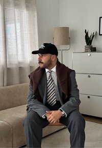 Costume gris avec un châle marron drapé, cravate rayée et casquette noire. Assis sur un canapé beige avec une lampe moderne et une plante en pot en arrière-plan.