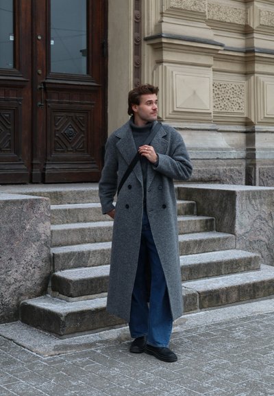 Abrigo de lana gris de doble botonadura con solapas anchas, combinado con jeans acampanados azul oscuro y zapatos negros, de pie en unos escalones de piedra.