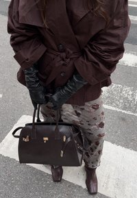 Trench coat marrone con maniche oversize, guanti in pelle nera, pantaloni a fantasia e una borsa a mano strutturata in pelle marrone con dettagli dorati.