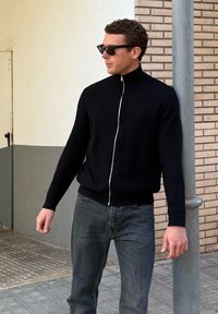 Homme portant un pull zippé noir et des lunettes de soleil, appuyé contre un poteau métallique sur un trottoir en ville.