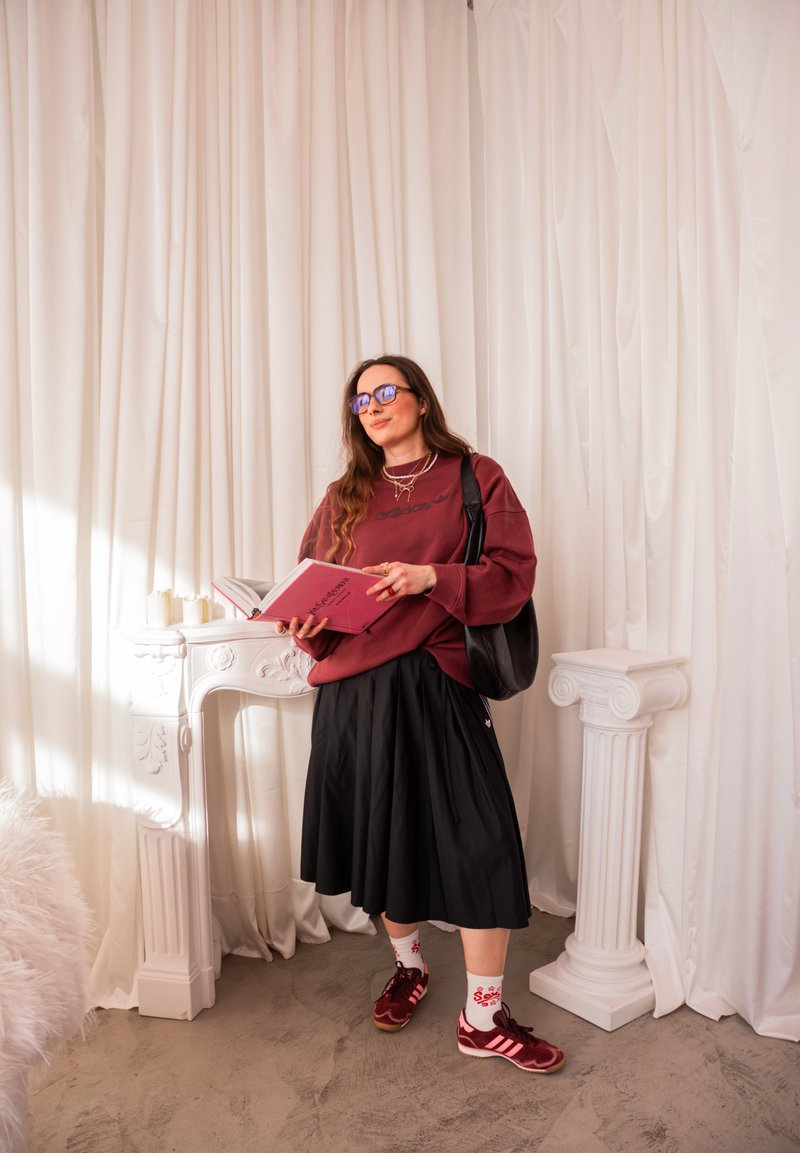 Jeune femme portant des lunettes, un sweat-shirt bordeaux, une jupe noire et des baskets, lisant un livre rose près de colonnes décoratives blanches et de rideaux drapés.