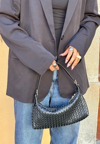 Mala de mão em couro preto trançado com forma curva e alça curta, segurada numa mão, combinada com uma blusa preta e um blazer oversized.