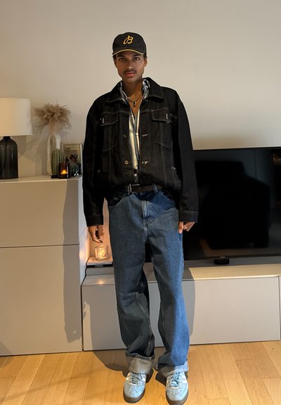 Chaqueta de mezclilla negra sobre una camisa a rayas, jeans azules con los dobladillos remangados y zapatillas deportivas azul claro, llevadas con una gorra gris con un logotipo amarillo.