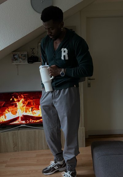 Cardigan verde oscuro con letras blancas, pantalones grises de corte relajado, sosteniendo una taza blanca, con un televisor mostrando un fuego al fondo.