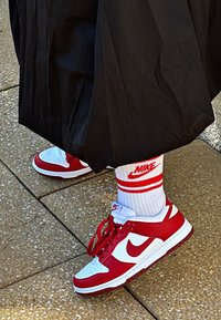 Sneakers Nike Dunk rossi e bianchi con punta rotonda e perforazioni, abbinate a calzini bianchi con righe rosse e logo, indossati sotto un indumento nero.