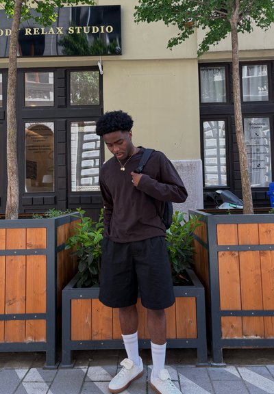 Camisa negra de manga larga, pantalones cortos negros, calcetines blancos y zapatillas claras. De pie frente a jardineras de madera con vegetación, cerca de una tienda.