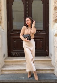 Bežani pleteni pulover s gumbima, kremasti satenski suknja i bež čizme do gležnja. Drži crnu torbu s zlatnim detaljem. Pozadina s smeškim drvenim vratima.