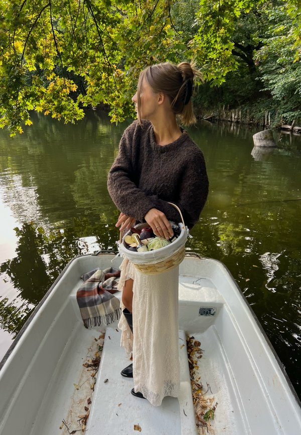Femme avec un panier de fruits debout sur une petite barque dans une eau calme, entourée d'arbres verts aux branches pendantes.