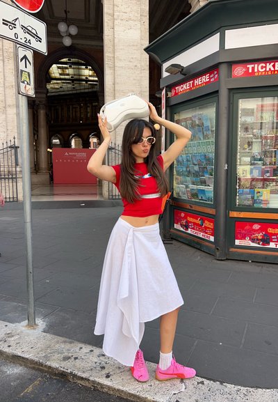 Una falda blanca cae de manera asimétrica sobre un top rojo corto, combinado con zapatillas rosas. Se sostiene un bolso blanco por encima, con gafas de sol que realzan el look.