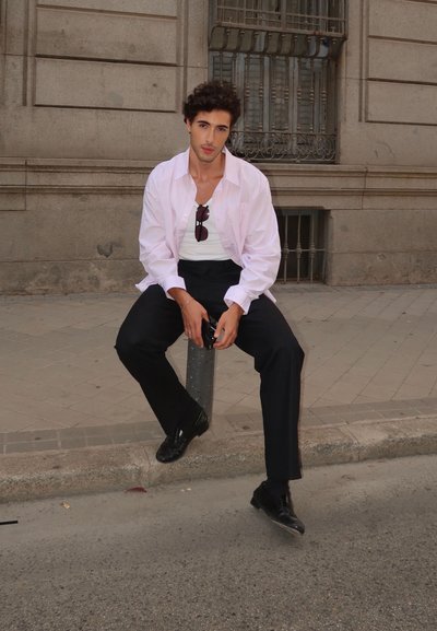 Camisa blanca de manga larga sobre una camiseta blanca ajustada, combinada con pantalones negros y zapatos negros brillantes, sentado en un poste gris contra una pared de piedra.