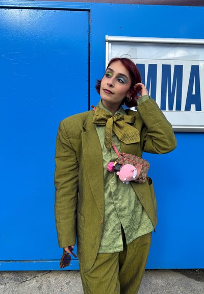 Persona con cabello corto y rojo, vistiendo un traje verde oliva de gran tamaño y una camisa estampada, sosteniendo unas gafas de sol, de pie frente a una pared azul con un cartel parcial.