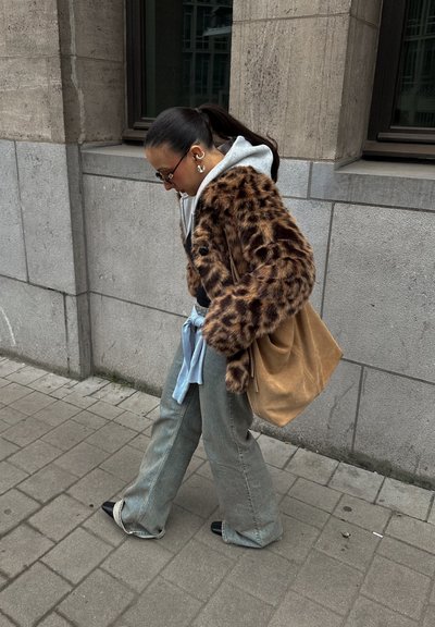 Mujer con coleta que lleva un abrigo con estampado de leopardo, vaqueros grises y zapatos negros caminando por una acera urbana junto a una pared de edificio de cemento.