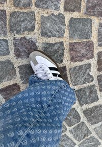 Białe sportowe buty z czarnymi paskami i szarymi zamszowymi akcentami, w połączeniu z wzorzystymi niebieskimi dżinsami, na tle kostki brukowej.