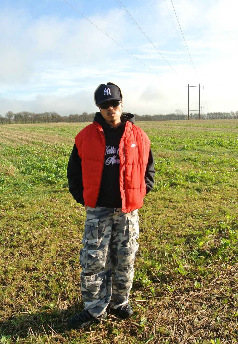 Κόκκινο γιλέκο πάνω από μαύρη κουκούλα, παραλλαγές φορτηγών παντελονιών. Μαύρο καπέλο και γυαλιά ηλίου, στέκεται σε ένα πράσινο χωράφι με ορατές γραμμές ρεύματος.