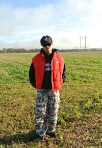 Κόκκινο γιλέκο πάνω από μαύρη κουκούλα, παραλλαγές φορτηγών παντελονιών. Μαύρο καπέλο και γυαλιά ηλίου, στέκεται σε ένα πράσινο χωράφι με ορατές γραμμές ρεύματος.