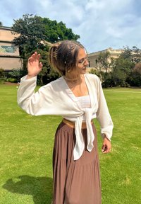 Młoda kobieta z warkoczami i okularami przeciwsłonecznymi, ubrana w białą bluzkę z wiązaniem na przodzie i brązową spódnicę, stoi na zielonym trawniku w świetle słonecznym.