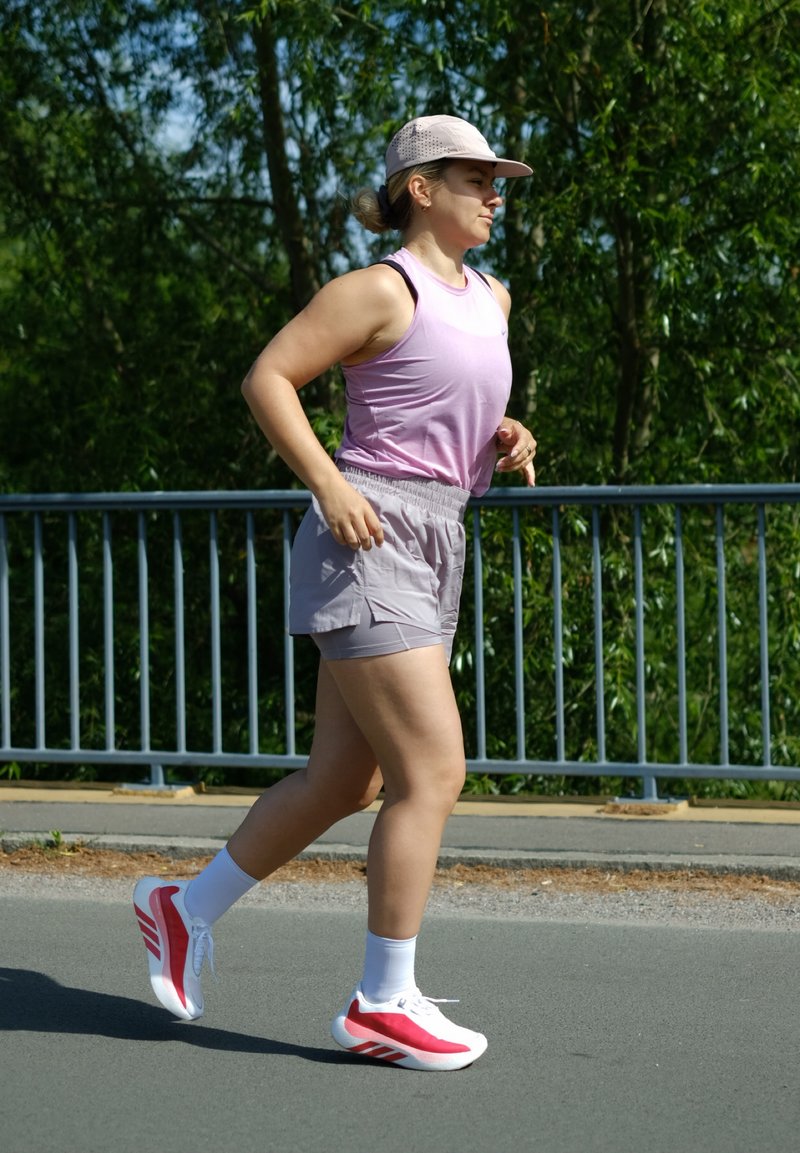 Femme courant à l'extérieur portant un débardeur rose, un short gris clair, une casquette rose, des chaussettes blanches et des chaussures de course rouges et blanches, près d'une rambarde métallique.