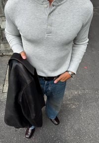 Camiseta de manga larga henley de canalé gris con tres botones, combinada con jeans azules y una chaqueta de cuero negra sostenida en una mano. Zapatos marrones visibles.