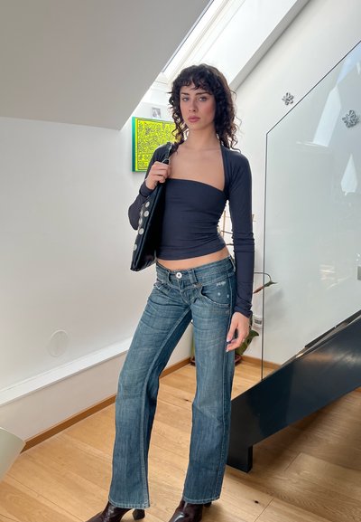 Cárdigan negro de manga larga sobre una blusa ajustada oscura, combinado con jeans de mezclilla de campana de un azul claro. Modelo sosteniendo un bolso negro, de pie en interiores.