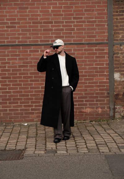 Abrigo negro de doble botonadura sobre un suéter crema, pantalones gris a rayas y calzado negro. Lleva una gorra de color claro y gafas de sol.