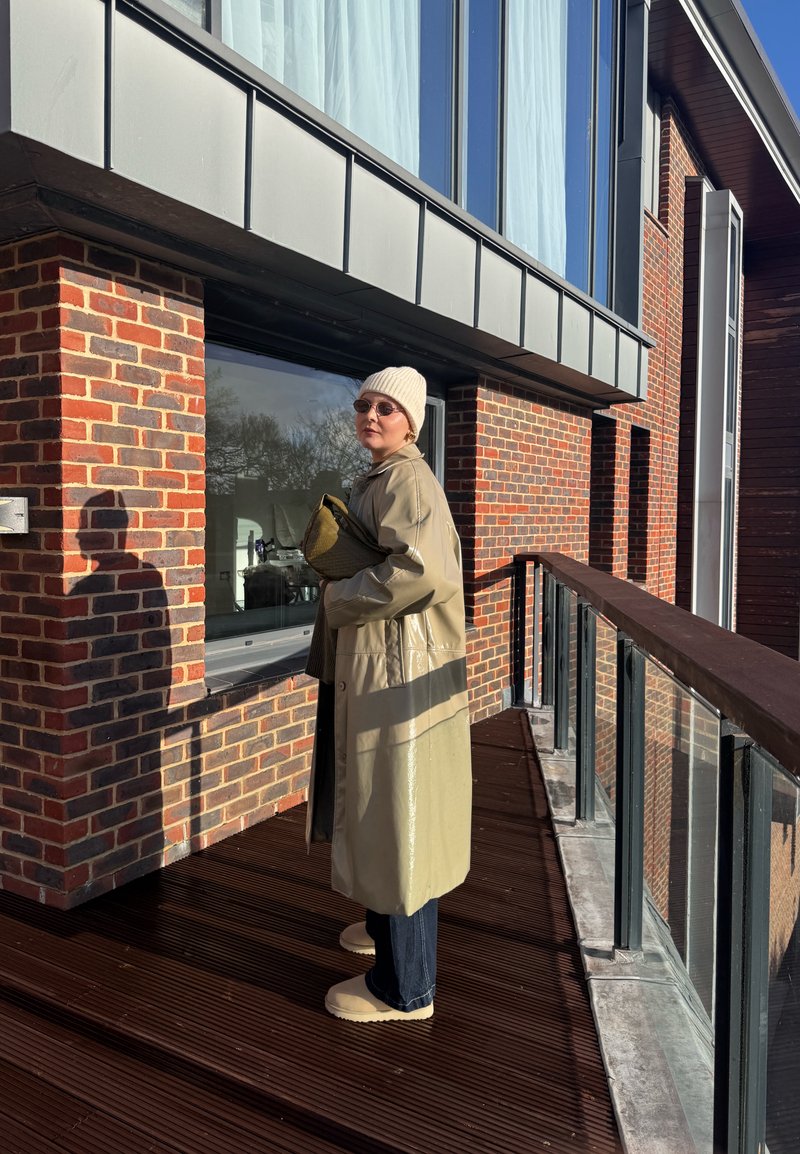 Personne portant un manteau beige, un bonnet, des lunettes de soleil et tenant un sac se tient sur un balcon en bois avec une rambarde en verre à côté d'un bâtiment en briques.