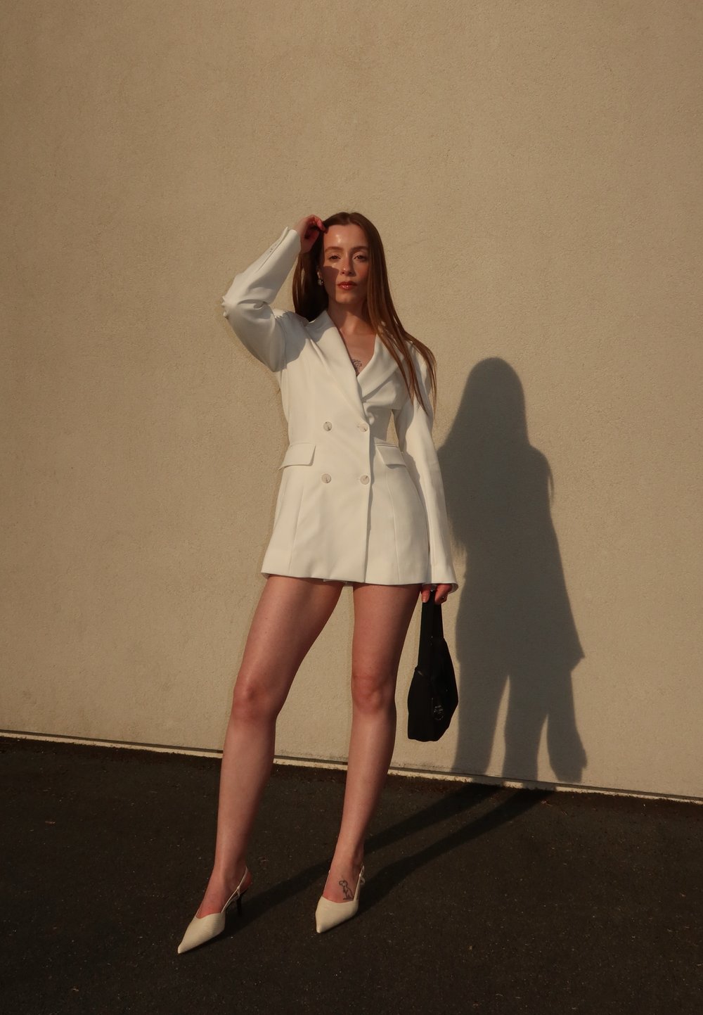 Hvit dobbeltradet jakke med strukturerte skuldre, kombinert med kort hem. Svart håndveske og kremfargede spisse hæler. Jevn tekstur.