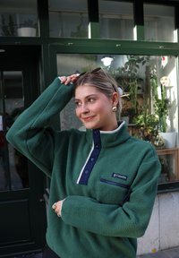 Pulóver de forro polar verde con cuello y botones azul marino, bolsillo con cremallera y logo. Textura suave, diseño de media cremallera y ajuste relajado.